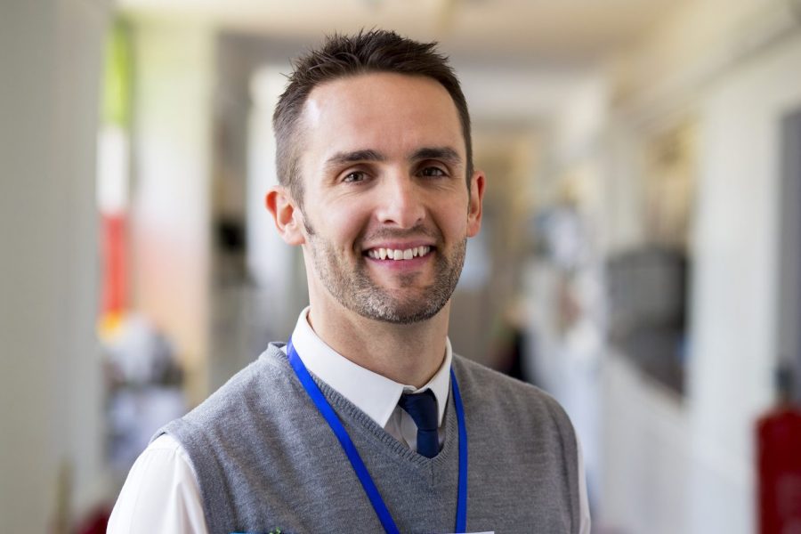 A happy male teacher dressed smartly and smiling in a school corridor. He is holding folders and a digital tablet.