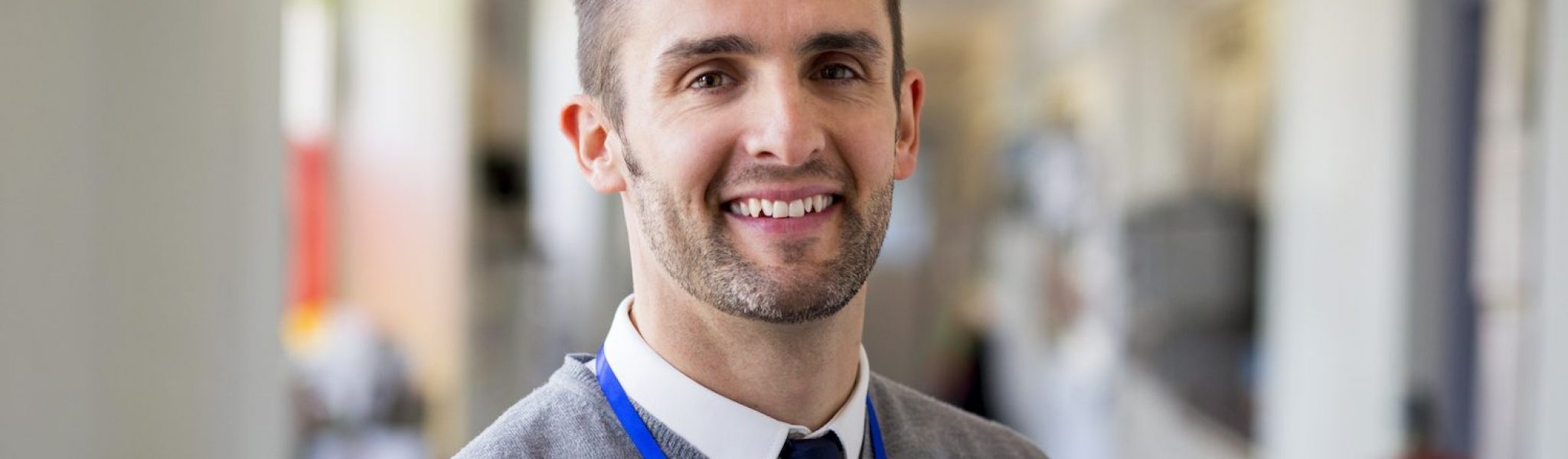 A happy male teacher dressed smartly and smiling in a school corridor. He is holding folders and a digital tablet.