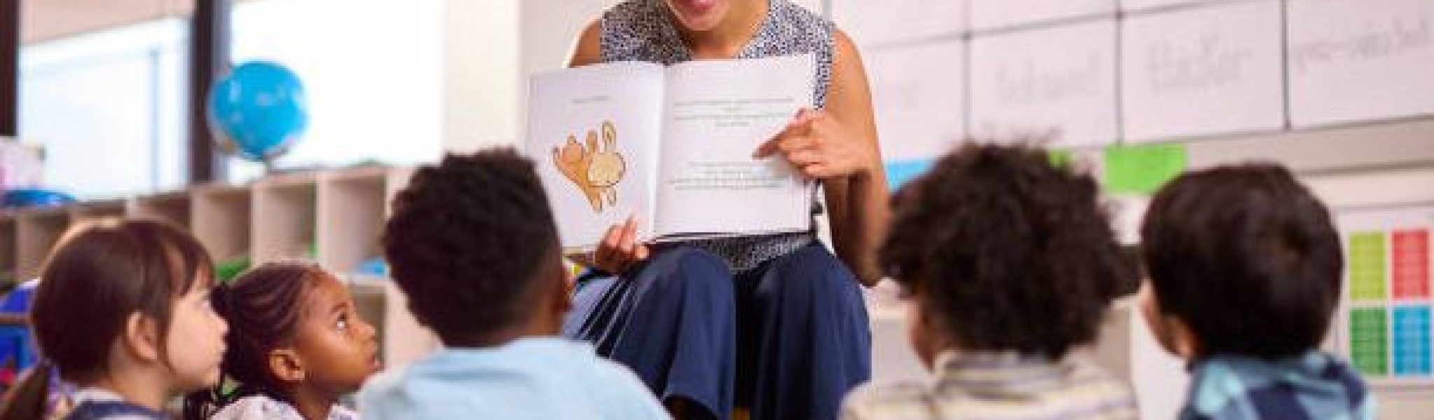 Female Teacher Reads To Multi-Cultural Elementary School Pupils Sitting On Floor In Class At School