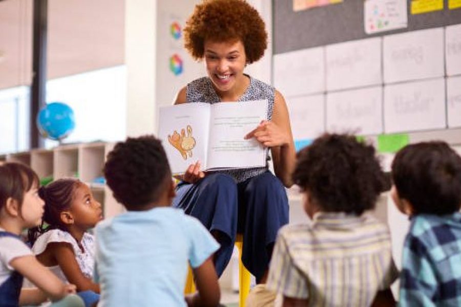 Female Teacher Reads To Multi-Cultural Elementary School Pupils Sitting On Floor In Class At School
