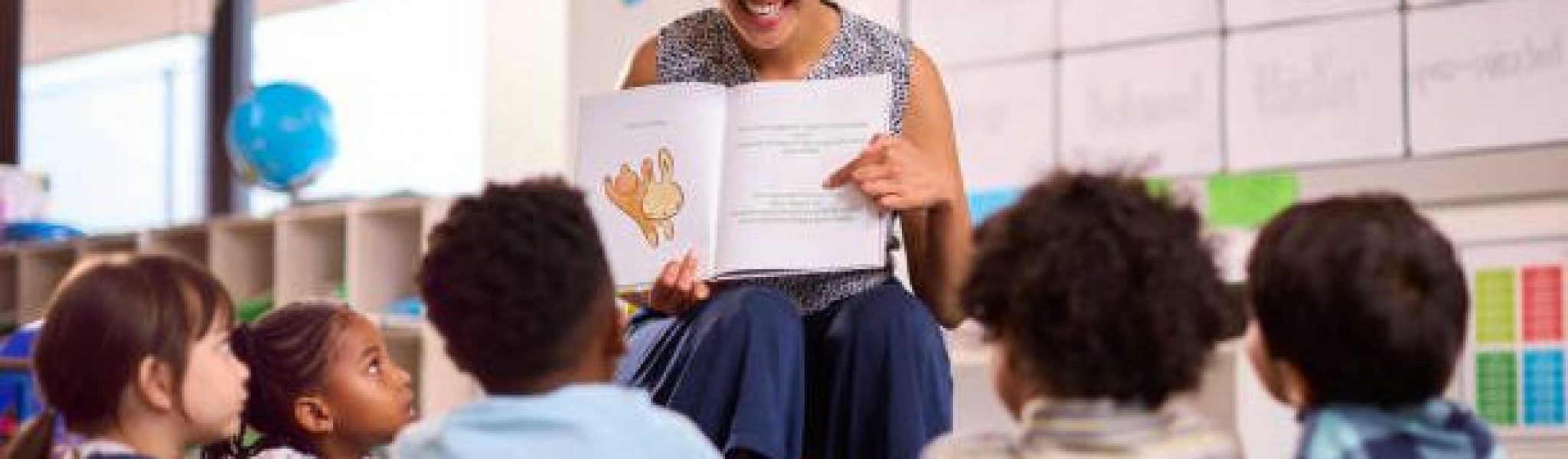 Female Teacher Reads To Multi-Cultural Elementary School Pupils Sitting On Floor In Class At School